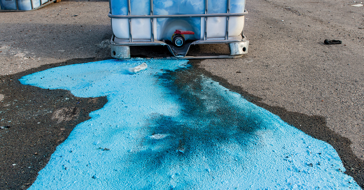 Image of a white plastic bulk container that has leaked a bright blue liquid onto a large area on the ground.