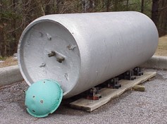 Image of a large silver cylinder laying horizontal on a base with a concave end and a small turquoise cap propped against the end.