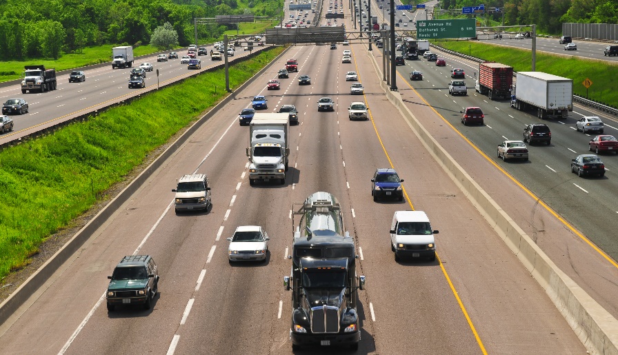 Image of a multilane divided highway, containing different passenger automobiles and large trucks with trailers.