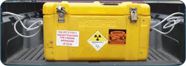 Image of a yellow radioactive materials package in the back of a truck bed. It is a sealed yellow rectangular container with two latches and a radioactive placard sticker on the front.